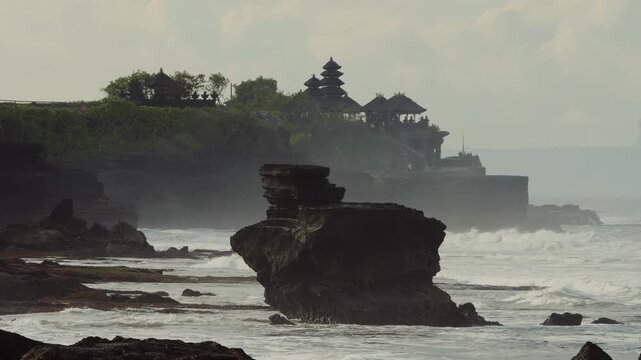 Tanah lot temple,Moody Coastline With Rolling Waves, Overcast Sky And Dramatic Clouds, Rocky Foreground With Distant Temple Pagoda, Cinematic Documentary Travel Vibe And Textured Seascape.