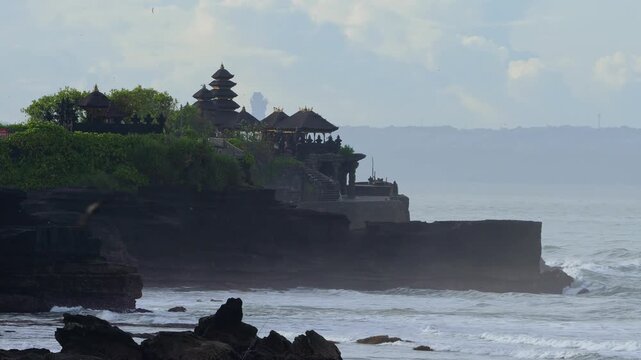 Pagoda Peeking Over Green Foliage On Bali Coastline, Distant Temple Silhouette Framed By Shrubs, Calm Blue Sky Background, Quiet Lookout Mood Evoking Pilgrimage And Cultural Discovery,Tanah Lot temple