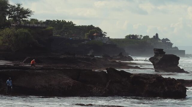 Rock Formation Framed By Surf,Towering Sea Stack With Pounding Waves,Mist Veil And Fierce Spray,Distant Temple Pagoda Tanah Lot On Cliff, Moody Light, Geology And Erosion Focus For Travel Documentary.