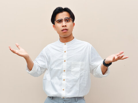 Confused young Asian man shrugging shoulders with open palms gesture against plain background.