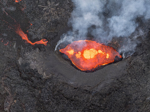 Aerial view of a volcanic eruption with glowing molten lava flowing from a crater surrounded by dark solidified volcanic rock and rising smoke in Iceland.