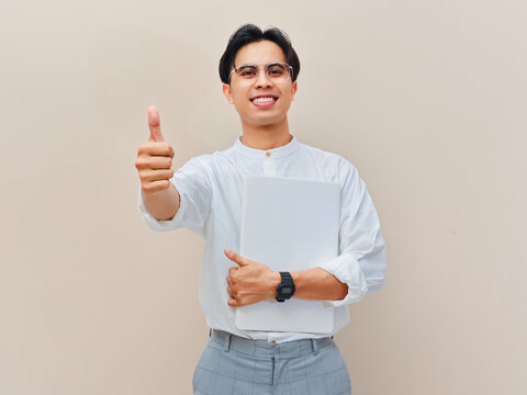 Cheerful young Asian professional or entrepreneur holding a closed laptop and giving a thumbs up gesture of approval.