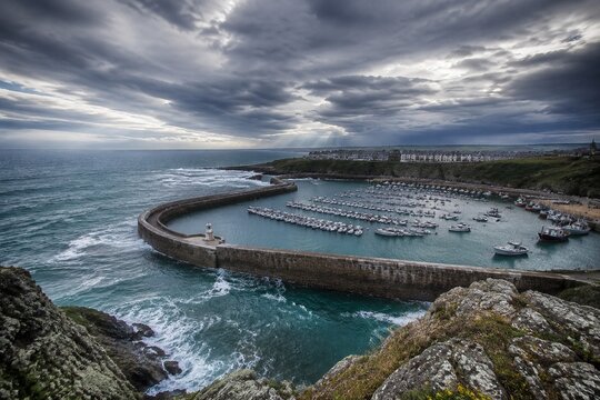 Aerial view from a cliff showing orderly ships in a harbor contrasted with rough seas.