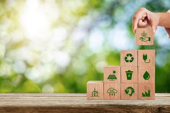 A hand placing a wooden block on a pyramid of cubes featuring environmental symbols for carbon reduction, recycling and renewable energy in a natural green setting.