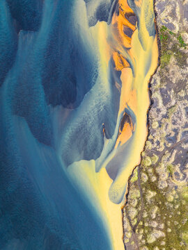 Aerial view of the vibrant glacial river delta featuring striking blue and yellow sediment patterns flowing through dark volcanic sand and mossy terrain in Iceland.