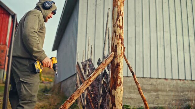 A Man is Using a Power Drill to Work on a Wooden Post as Part of an Outdoor Fence Construction - Medium Shot