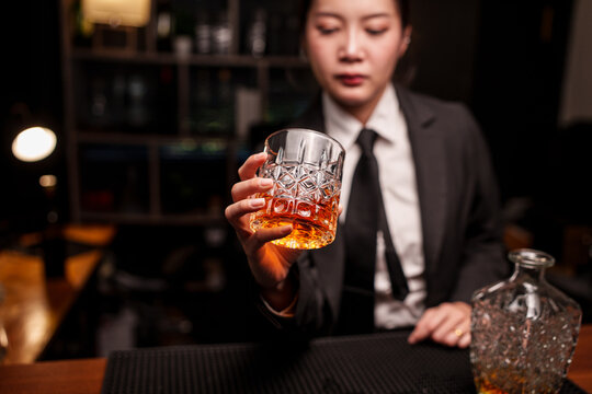 Woman bartender in a suit is handing a glass of whiskey across the bar counter.
