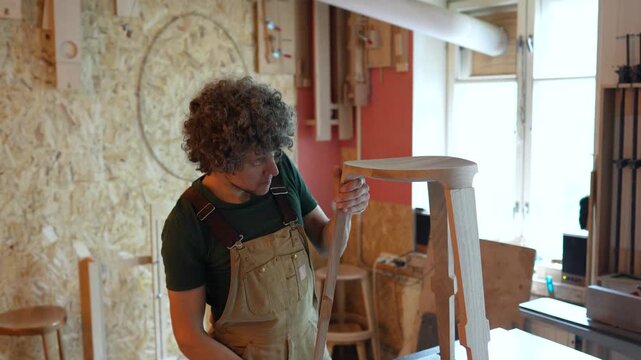 Private wooden furniture manufacture, man carpenter working in workshop with wooden chair, attentive male woodworker making adjustments to half-finished stool legs, fitting and using instruments.