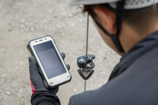 測量機器を確認しながらハンディ端末を操作する建設作業員。ヘルメット姿の現場作業