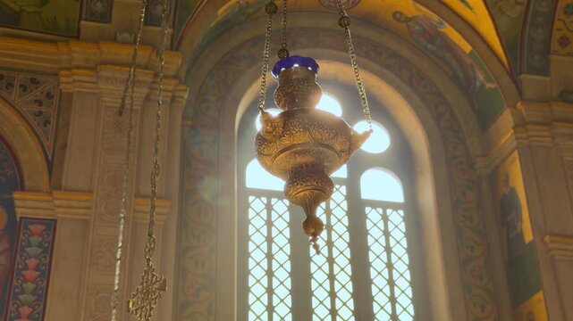 Ornate golden oil lamp hanging on chains inside orthodox church with painted arches and window light, sacred temple interior, religious tradition, spiritual heritage concept.
