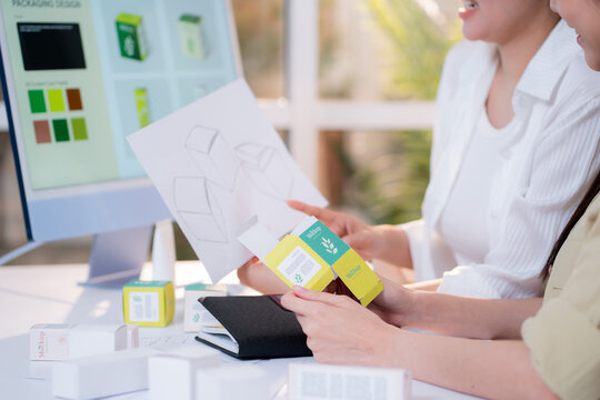 Two asian woman graphic designers analyzing product package prototype, reviewing paper sketch at home office, comparing physical box mockup with computer screen display with artificial intelligence.