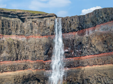 Aerial view of Hengifoss waterfall cascading down dramatic layered basalt and red clay cliffs in Iceland.