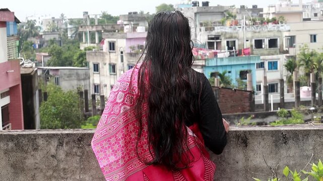 Back view of a Bangladeshi woman with long wet black hair in pink saree looks at the city buildings from her terrace during daytime. The lady displays deep loneliness and sadness.