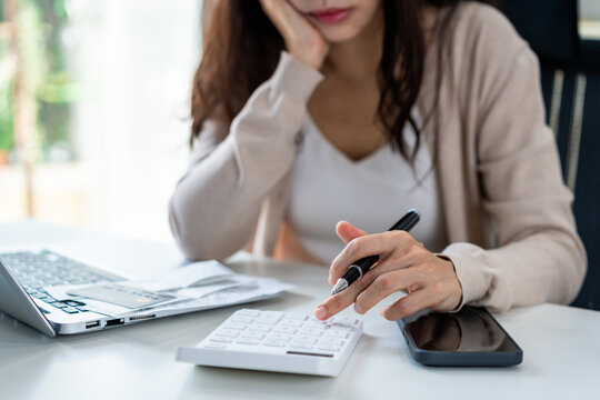 A pensive young woman uses a calculator with credit cards and a smartphone, deeply focused on her desk as she carefully calculates her monthly household expenses and creates a budget