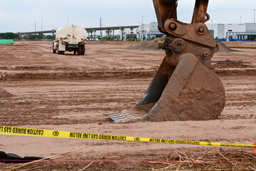 Excavator bucket in the foreground of a large commercial development site with a water truck in the background, representing earthmoving, site preparation, and infrastructure construction activity © Eduardo Barraza