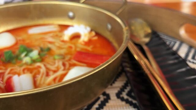 Close up slow motion top view of delicious spicy Tom Yam noodle soup with crab sticks, fish balls, and quail eggs served in a gold pot on a rustic wooden tray
