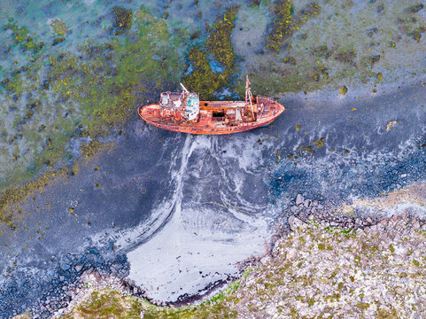 Aerial view of a rusted shipwreck stranded on a dark volcanic sand beach with mossy rocks and shallow coastal waters in Iceland.