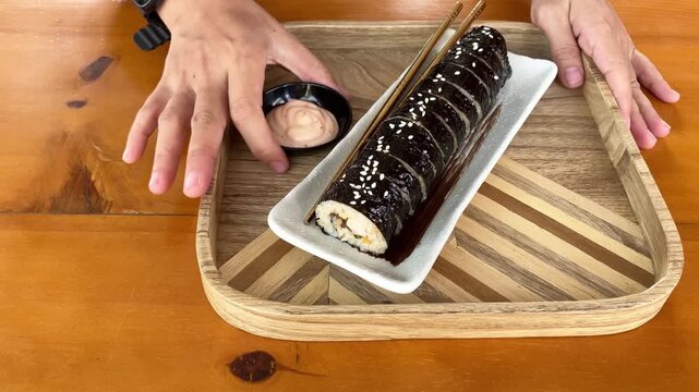 Slow motion view of a person's hands serving a small bowl of sauce next to a rectangular plate of Korean gimbap seaweed sushi roll with gold chopsticks on a patterned wooden tray