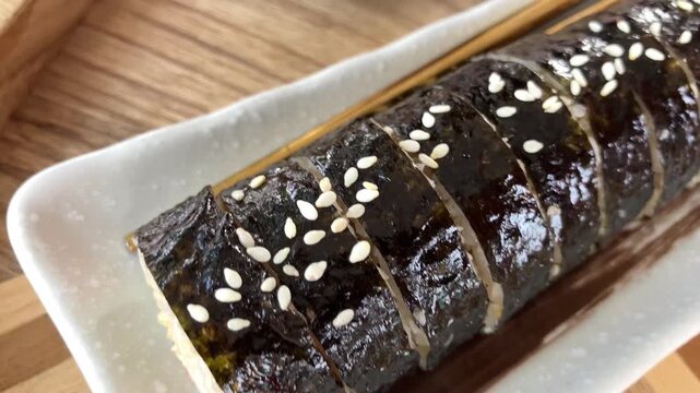 Close up macro view of delicious Korean gimbap seaweed sushi rolls sprinkled with sesame seeds, served on a white rectangular plate with chopsticks resting on a patterned wooden tray