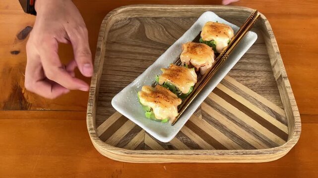 Slow motion view of a person's hand serving a rectangular plate of delicious dim sum shumai topped with creamy torched mentai sauce onto a wooden tray on a table