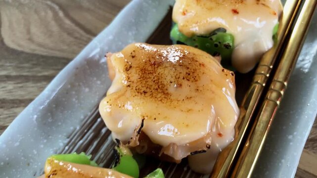 Close up macro view of delicious dim sum shumai topped with creamy torched mentai sauce, served on a rectangular plate with gold chopsticks