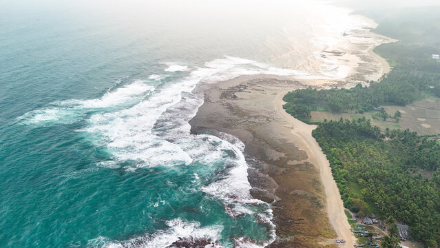 Aerial view of a rugged coastline with turquoise waves crashing against the rocky shore, bordered by a lush palm tree forest and sandy beach in Indonesia.