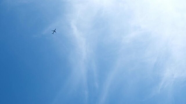 An airplane soars through a clear blue sky with wispy clouds. The aircraft is small and appears high above the ground, emphasizing the vastness of the sky.
