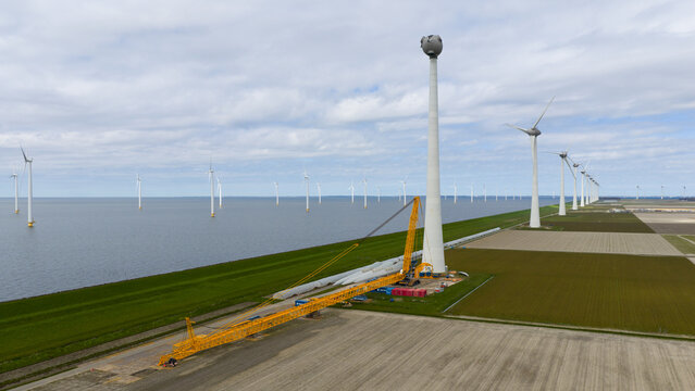 Aerial view of a large yellow crane performing maintenance on a wind turbine tower, with rows of turbines in the water and agricultural fields in Espel, Flevoland, Netherlands.