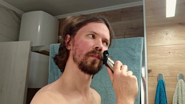 A man with long brown hair shaves his beard using an electric razor in a bathroom. He has a light stubble and is shirtless.
