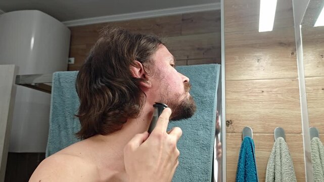 A man with long brown hair shaves his beard using an electric razor in a bathroom. He is shirtless and focused on grooming.