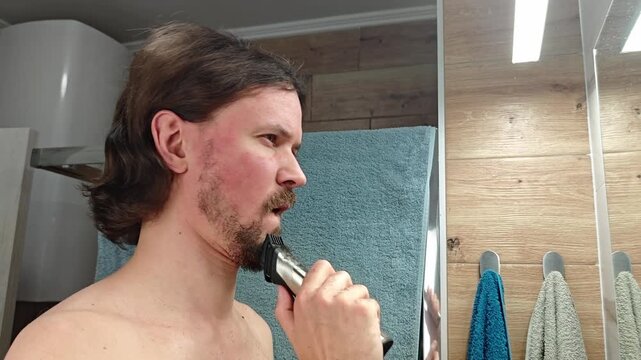 A young Caucasian man with long brown hair shaves his beard using an electric razor in a bathroom setting. Towels hang in the background.