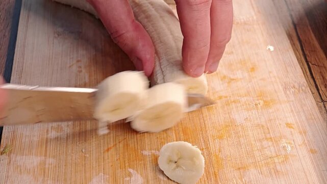 A person slices a ripe banana into rounds with a sharp knife on a wooden cutting board.