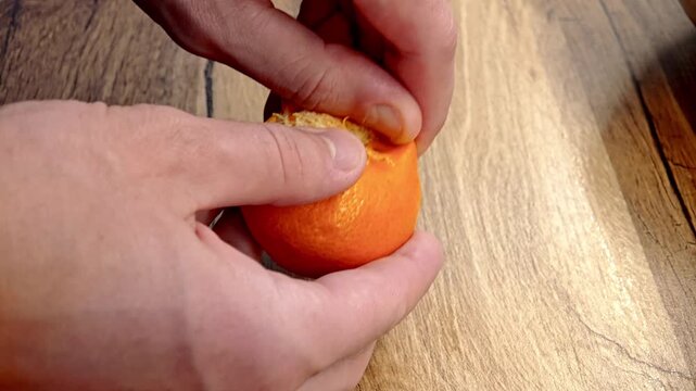 A person peels a mandarin on a wooden surface. The fruit is bright orange and round, with the peeling process clearly visible.