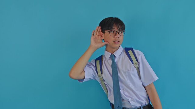 Asian high school student holding hand to ear gesturing listening closely or eavesdropping on blue background