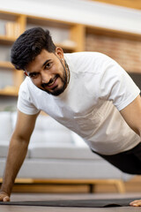 Fototapeta premium A young Indian man with a beard and a white t-shirt performs a push-up exercise on a mat in a modern living room setting