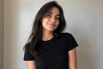 Fototapeta premium Young woman smiles softly against a plain wall. She wears a simple black t-shirt, long dark hair flows. Her gaze meets the viewer with calm, friendly warmth. Perfect for lifestyle, portrait