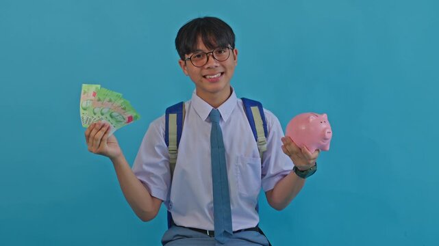 Happy Indonesian student holding rupiah cash banknotes and pink piggy bank on blue background