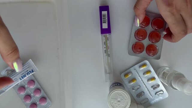 Woman's hands managing various medications like pills, capsules, nasal spray and thermometer inside a white container for organizing a home first aid kit