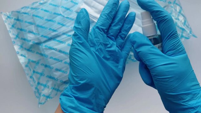 Person in blue nitrile gloves disinfecting hands with antiseptic spray over a package of new disposable medical masks, ensuring health safety and hygiene during a pandemic