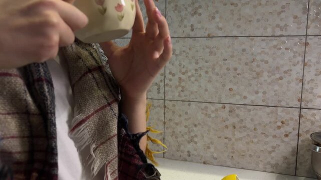 Woman with a plaid scarf enjoying a warm cup of herbal tea with lemon, relaxing at home in the kitchen and drinking a hot beverage to get warm and cozy on a cold day