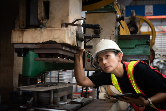 Young female factory worker in safety helmet and reflective vest inspects industrial press machine workwear factory industry with focused expression and tablet in hand