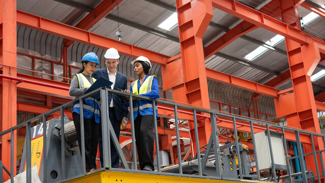 Three factory supervisors wearing workwear and helmets inspect machinery on elevated platform in large industrial warehouse with orange steel beams conveying focus and collaboration