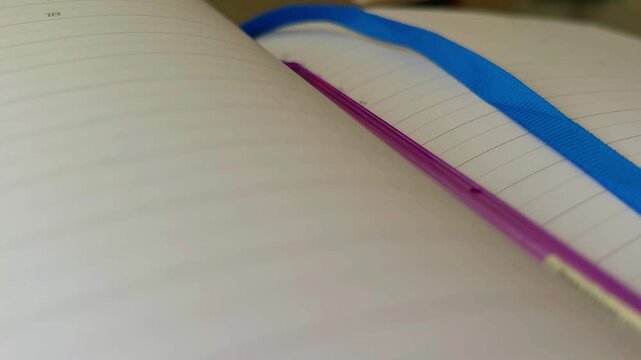 Flipping through a personal lined diary, turning the blank pages of a notebook with a blue ribbon bookmark and a purple pen, representing studying, writing, or journaling