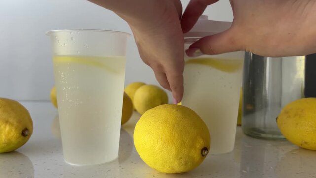 Hands closing a reusable plastic cup with a lid, preparing a refreshing cold homemade lemonade with ice and lemon slices on a bright kitchen counter with fresh lemons