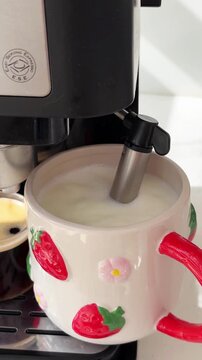 Close up view of a professional espresso machine with a steam wand frothing milk inside a large. Cute ceramic mug decorated with strawberries