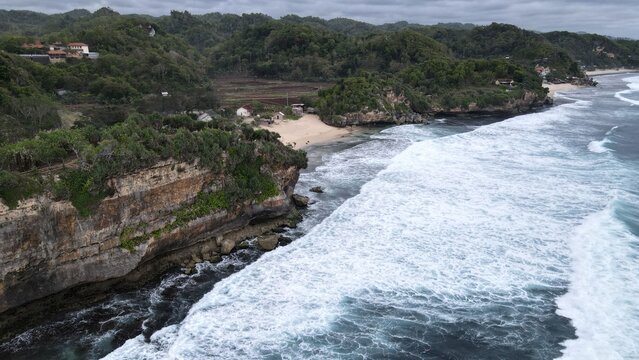 Aerial view of Indrayanti Beach with rugged limestone cliffs, white sand, crashing waves, and lush green vegetation in Kecamatan Tepus, Daerah Istimewa Yogya, Indonesia.