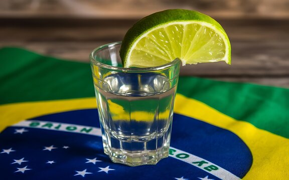 Small glass of clear brazilian cachaca spirit with a fresh lime wedge garnish resting on a colorful brazilian flag background