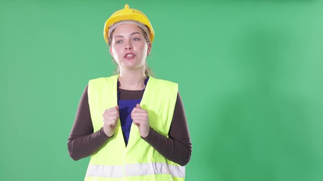 Portrait of young girl worker dressed in reflective vest and hard hat talking, actively gesturing and inviting or giving examples against green screen background 