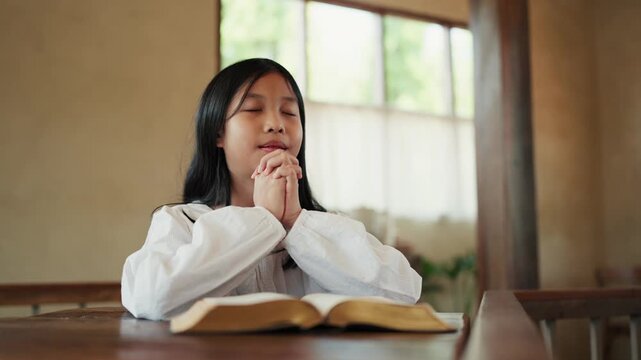 Child pray concept. Happy Asian girl with folded hands to prayer with bible and praise worship in church. kid praying to god. Christianity, spirituality, religion and faith.