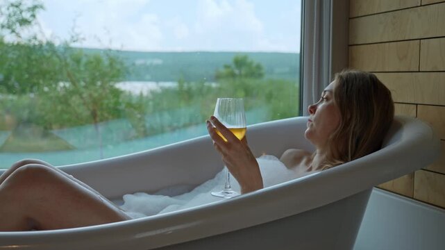 A woman unwinds in a luxurious bubble bath, holding a glass of wine, as she enjoys a magnificent daytime view of a lake and green landscape from a large window.